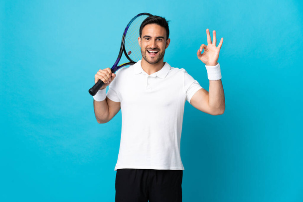 Genç yakışıklı adam mavi arka planda izole edilmiş tenis oynuyor ve tamam işareti yapıyor. - Fotoğraf, Görsel