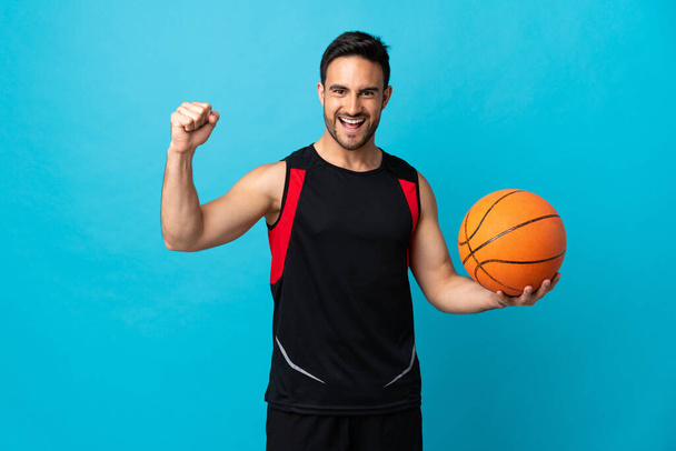 Fiatal jóképű férfi elszigetelt kék háttér játszik kosárlabda - Fotó, kép
