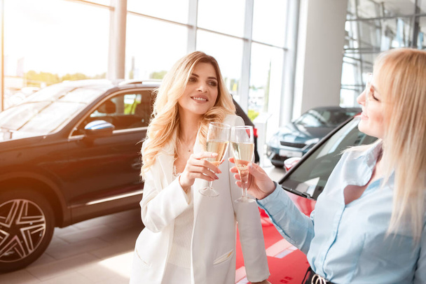 Two girlfriends at a car dealership rejoice in buying a new car with glasses of champagne in their hands - Фото, зображення