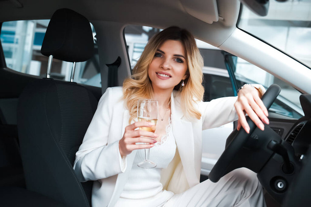 Geschäftsfrau mit einem Glas Champagner sitzt in ihrem neuen Auto in einem Autohaus. Mädchen feiert den Kauf eines neuen Autos - Foto, Bild