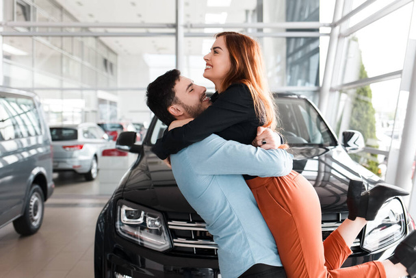 Mies halaa tyttöystäväänsä autokaupassa ostamansa auton taustalla. - Valokuva, kuva