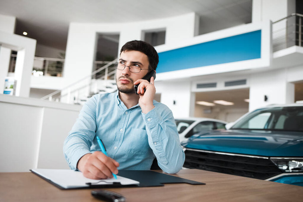 Pensive car dealership employee talking on the phone and writing - Photo, Image