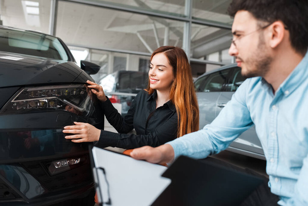 Venditore mostra l'auto ad un giovane cliente di una concessionaria di auto. Ragazza sceglie una nuova auto
 - Foto, immagini