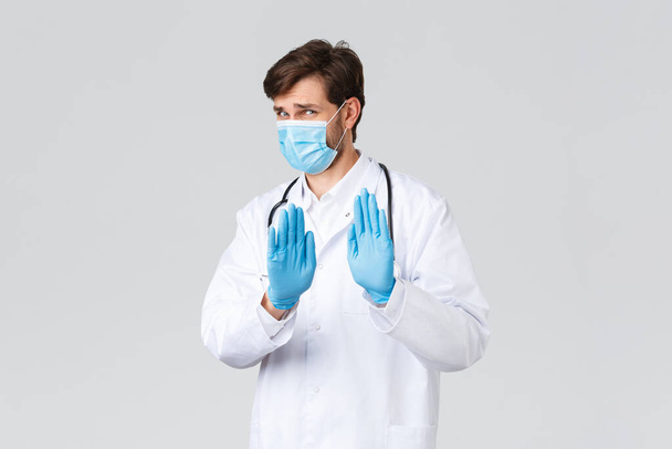 Hospital, healthcare workers, covid-19 treatment concept. Oh no thank you, step back. Reluctant and displeased doctor in scrubs, medical mask and gloves, waving hands in rejection or refusal - Photo, Image