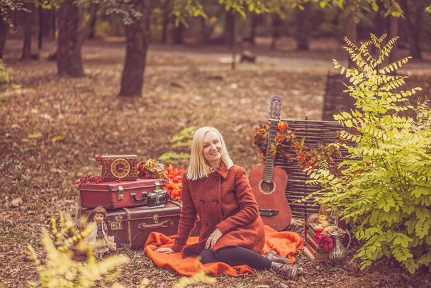 Photoshoot dans le parc avec des décorations. Une jeune femme blonde dans un manteau d'automne orange est assise sur un plaid orange avec un sourire sur son visage. - Photo, image