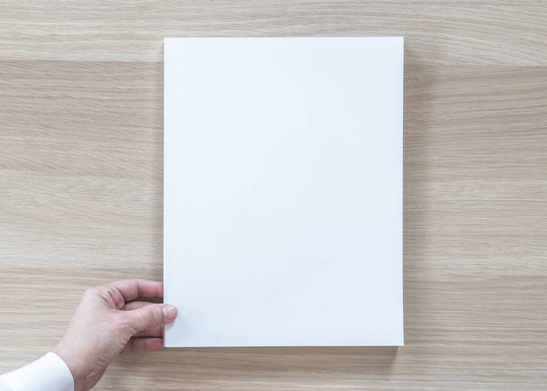 Blanco A4 formaat boek omslag model bovenaanzicht in de hand van de zakenman met pagina voorzijde op wit oppervlak plat lag op houten tafel - Foto, afbeelding