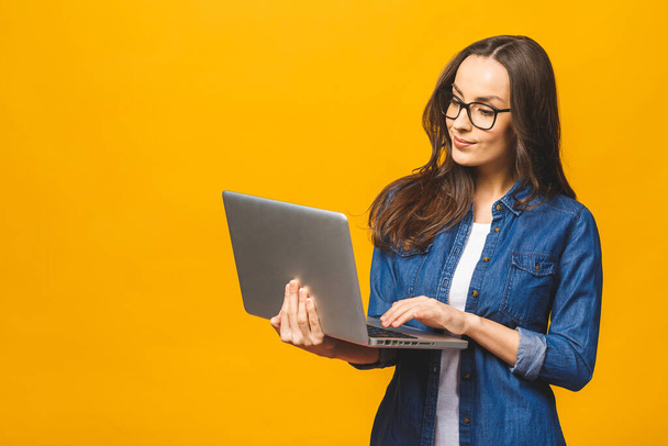 Jonge vrolijke glimlachende vrouw in casual kleding met laptop en het verzenden van e-mail naar haar beste vriend. Geïsoleerd tegen gele achtergrond.  - Foto, afbeelding
