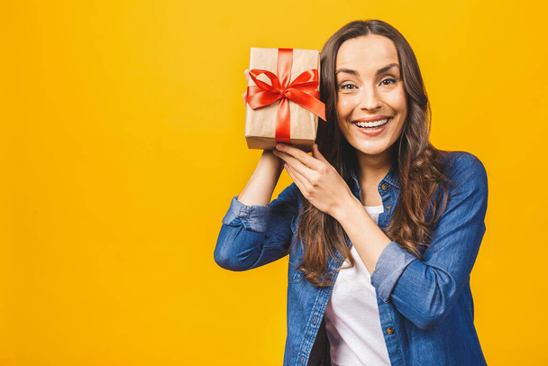 Heureuse femme brune dans une boîte cadeau tenue décontractée et regardant la caméra tout en appréciant sur fond jaune
. - Photo, image