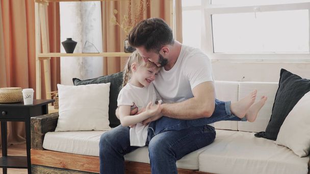 Любящий отец обнимает свою дочь на белом диване. Счастливые ребенок и папа чувствуют радость. Красивый папа и его милая маленькая девочка обнимаются, делятся любовью с отцом
 - Фото, изображение