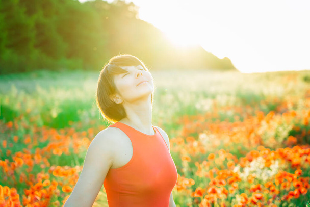 Close up happy young woman with closed eyes in red dress relaxing in red poppies flowers meadow at sunset. Celebrating freedom, positive emotions feeling life, peace of mind concept. Selective focus. - Photo, Image