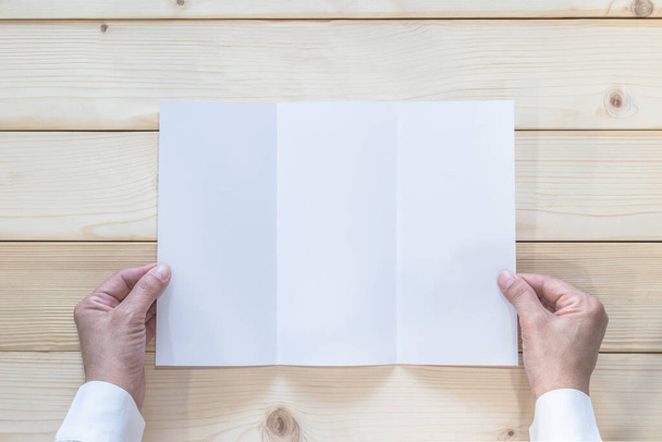 Λευκά φυλλάδια mock up τριών διπλωμένο φυλλάδιο Α4 χαρτί lay-out πρότυπο στο χέρι του επιχειρηματία στο ξύλινο τραπέζι από την κορυφή άποψη - Φωτογραφία, εικόνα