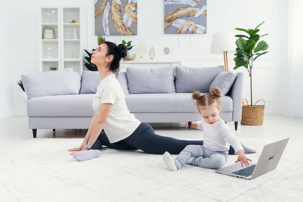 Lustiges Baby-Mädchen spielt mit Laptop, während ihre sportliche gesunde Mama zu Hause online Yoga trainiert. - Foto, Bild