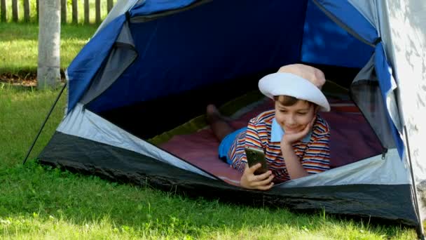 Il bambino si fa un selfie al telefono in una tenda. Ragazzino con un cappello si fotografa sul suo smartphone
. - Filmati, video