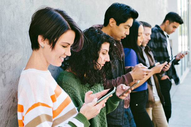 Группа молодых людей смотрит на свои мобильные телефоны, стоя у стены города - Millennial - Technology - Фото, изображение
