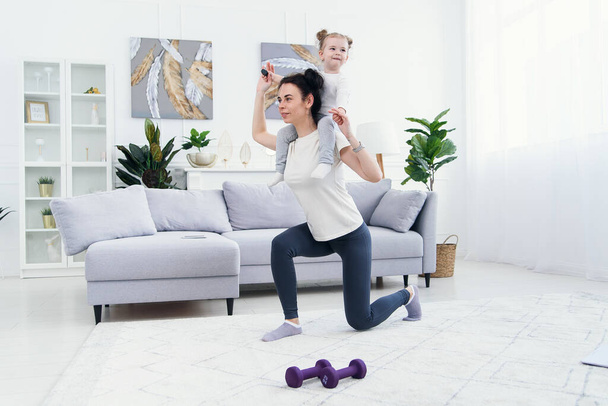 Beautiful mommy and charming little daughter are smiling while doing fitness exercises together at home. - Фото, зображення