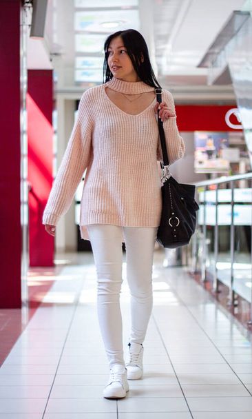 Kaunis hymyilevä tyttö kävelee ostoskeskuksessa. Hänellä on yllään kevyt villapaita ja valkoiset farkut ja musta laukku. - Valokuva, kuva