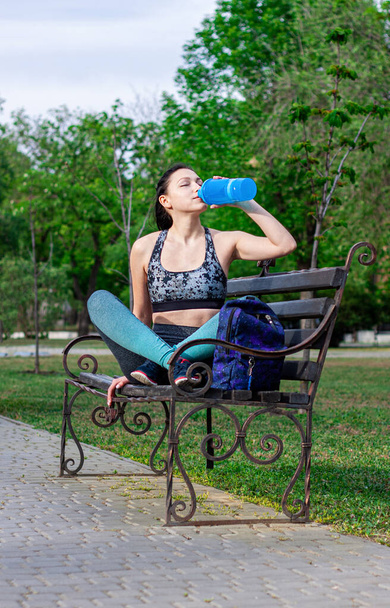 Beautiful  brunette girl with a backpack   is  sitting on the bench  in the park and drinks water aster training.  Concept of sport  - Photo, Image