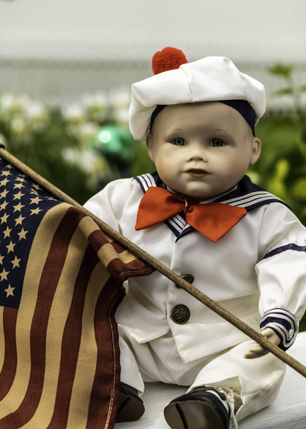 vintage small boy doll with glass eyes dressed in sailor costume vertical format outdoors holding american flag - Photo, Image