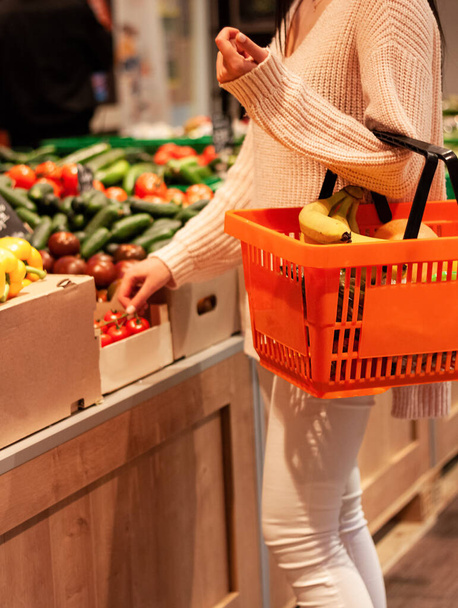 Mädchen nimmt einen Tomatenzweig und hält in einem Supermarkt einen orangefarbenen Korb voller Früchte in der linken Hand - Foto, Bild