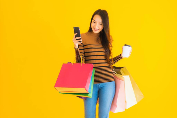 美しい若いアジアの女性は、黄色の隔離された背景にコーヒーカップとカラーショッピングバッグとスマートフォンや携帯電話を使用しています - 写真・画像