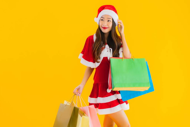 Porträt schöne junge asiatische Frau tragen Weihnachtskleidung Hut mit einer Menge Einkaufstasche auf gelbem Hintergrund isoliert - Foto, Bild