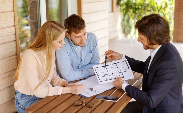 Real estate agent showing house plan to millennial couple at table on porch - Photo, Image