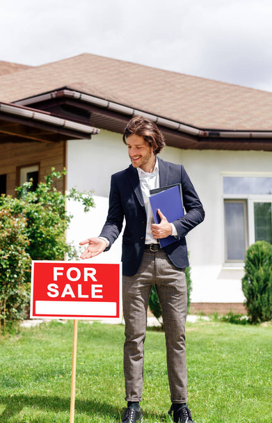Young real estate agent near beautiful house showing FOR SALE sign - Photo, Image