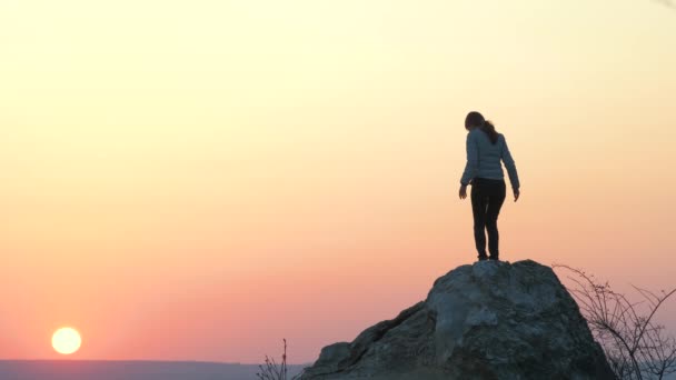 山の中で日没の大きな石の上に一人で登る女性ハイカーのシルエット。女性の観光客は夜の自然の中で高い岩の上に手を上げる。観光、旅行、健康的なライフスタイルの概念. - 映像、動画