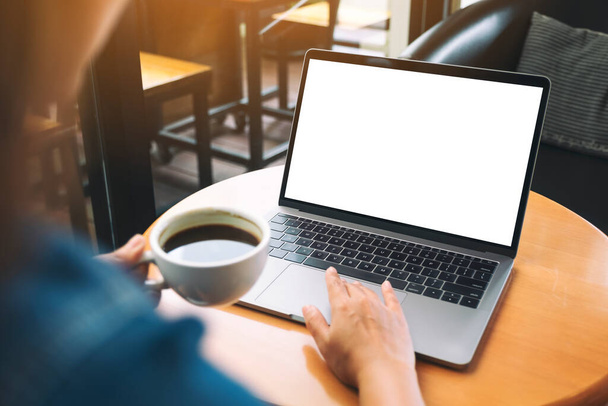 Mockup-Bild einer Hand, die beim Kaffeetrinken ein Laptop-Touchpad mit leerem weißen Desktop-Bildschirm benutzt und berührt - Foto, Bild