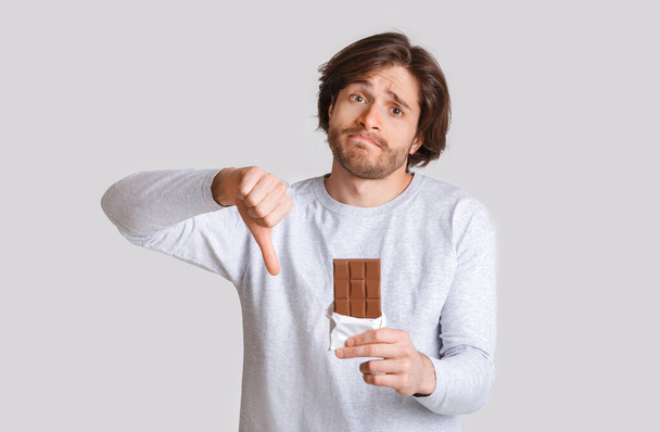 Diabetes and food allergies. Sad man shows thumb down and holds chocolate - Photo, Image