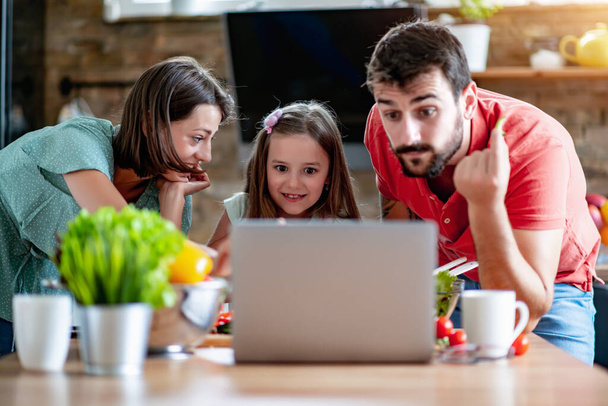 Cooking, food and home concept -mom, dad and child are cutting vegetables to make salad in the kitchen.They have fun and watching the recipe online. - 写真・画像