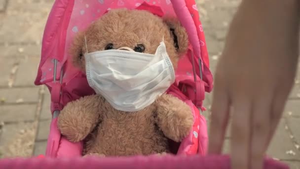 kid on street with his favorite toy in a protective mask. healthy childhood concept. little girl walks in park with a pram and a teddy bear in medical mask. child plays an epidemic and protects toy. - Video