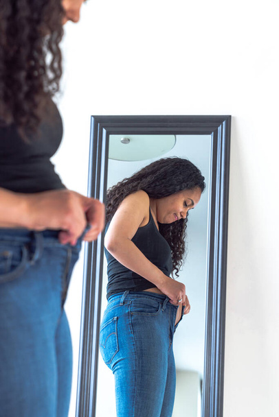 Beautiful African American woman and mother stands in front of a mirror and attempts to try to close her tight fitting blue jeans pants after postpartum diet and weight loss exercise. - Foto, immagini