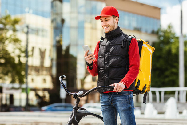 Mensajero usando teléfono celular entregando comida de restaurantes en bicicleta al aire libre
 - Foto, Imagen