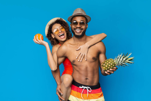 Sommerzeit. Fröhliches afrikanisches Paar in Badebekleidung mit tropischen Früchten in den Händen - Foto, Bild