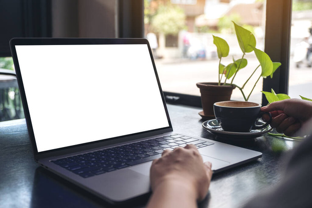 Imagem Mockup das mãos de uma mulher usando e tocando touchpad laptop com tela de desktop branca em branco enquanto bebe café na mesa de madeira no café
 - Foto, Imagem