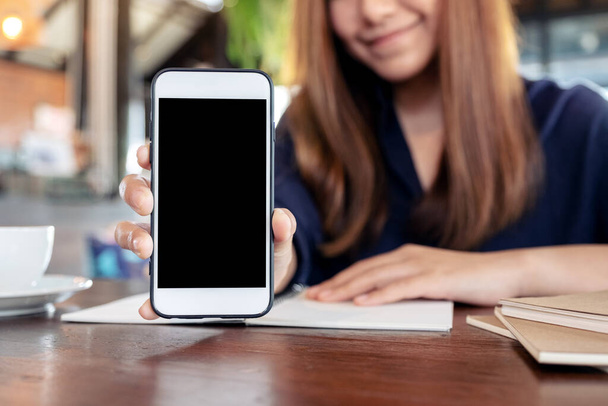 Макет азіатської жінки, що тримає і показує білий мобільний телефон з порожнім чорним екраном на столі під час роботи
  - Фото, зображення