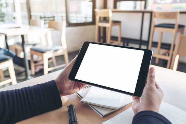 Mockup afbeelding van handen houden en het gebruik van zwarte tablet pc met blanco witte desktop scherm tijdens het werken aan notebooks op kantoor - Foto, afbeelding