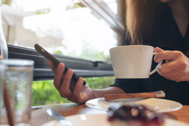 Κοντινή εικόνα μιας γυναίκας που κρατάει, χρησιμοποιεί και αγγίζει ένα έξυπνο τηλέφωνο ενώ πίνει καφέ - Φωτογραφία, εικόνα