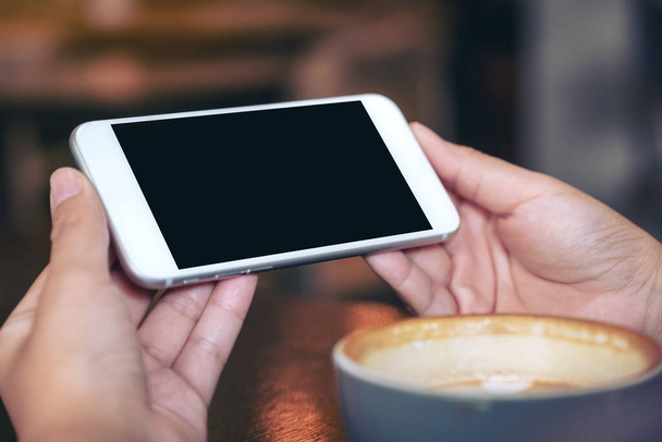 Mockup-Bild von Händen, die ein weißes Mobiltelefon mit schwarzem Bildschirm horizontal halten und verwenden, um mit Kaffeetasse auf Holztisch zuzusehen - Foto, Bild