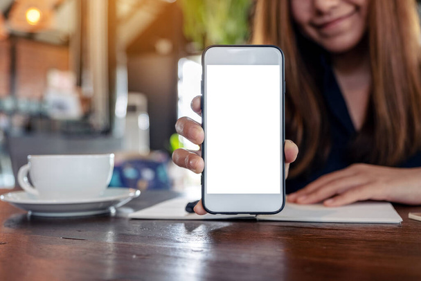 Mockup obraz asijské ženy drží a ukazuje bílý mobilní telefon s prázdnou stolní obrazovku s šálky kávy a notebooky na ročníku dřevěný stůl v kavárně - Fotografie, Obrázek