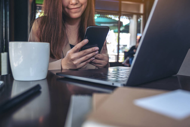 オフィスのテーブルの上にコーヒーカップでノートパソコンで作業しながら、スマートフォンを持ち、使用し、見ているアジアの女性のクローズアップイメージ - 写真・画像
