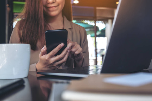 Nahaufnahme einer asiatischen Frau, die während der Arbeit am Laptop mit Kaffeetasse auf dem Tisch im Büro Smartphone hält, benutzt und anschaut - Foto, Bild