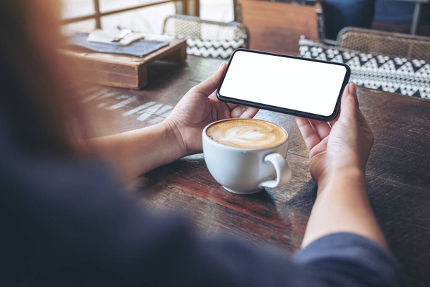 Mockup image of a woman holding and using a black mobile phone with blank screen horizontally for watching with coffee cup on wooden table - Photo, Image