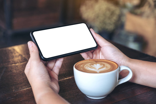Mockup-Bild von Händen, die ein schwarzes Mobiltelefon mit leerem Bildschirm horizontal halten und verwenden, um mit Kaffeetasse auf Holztisch zuzusehen - Foto, Bild