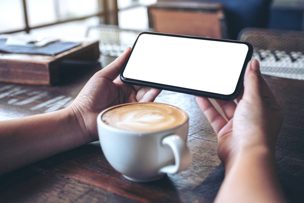 Образец изображения рук, держащих и использующих черный мобильный телефон с пустым экраном горизонтально для просмотра с чашкой кофе на деревянном столе
 - Фото, изображение