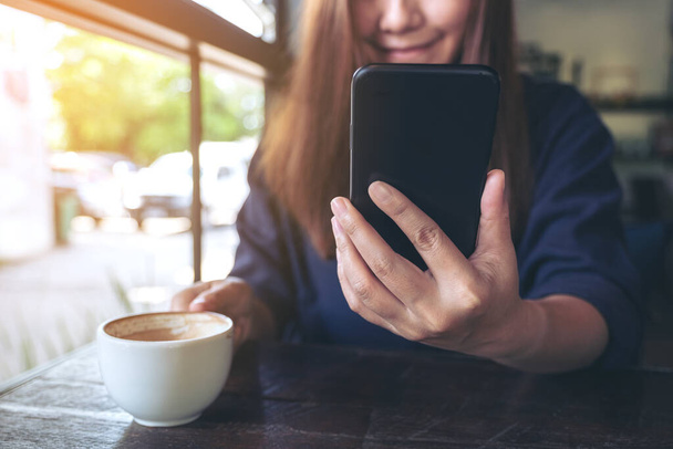 Primer plano imagen de una hermosa mujer asiática sosteniendo, usando y mirando el teléfono inteligente mientras bebe café en la cafetería - Foto, Imagen