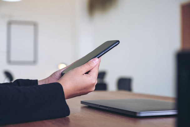 Жінка тримає і використовує чорний мобільний телефон з ноутбуком на дерев'яному столі в офісі
 - Фото, зображення