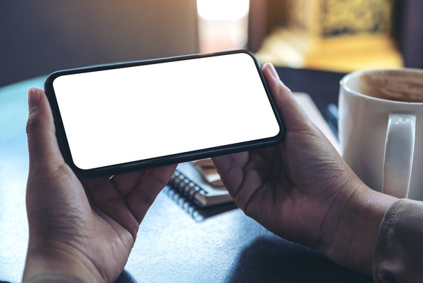 Mockup-Bild von Händen, die ein schwarzes Mobiltelefon mit leerem Bildschirm horizontal halten und verwenden, um mit Kaffeetasse und Notizbüchern auf dem Tisch zuzusehen - Foto, Bild