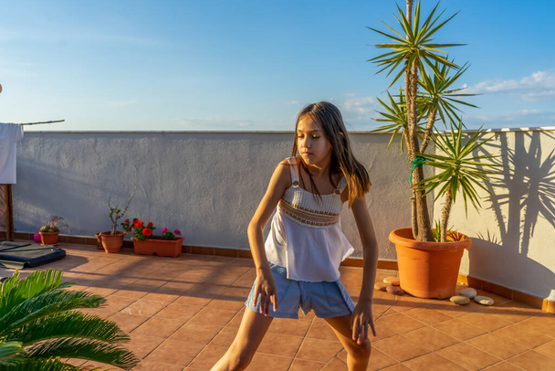 Linda niña vestida con una camiseta blanca bailando y saltando en la terraza de la casa al atardecer. Concepto divertido
 - Foto, imagen